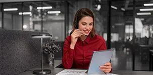 A customer support representative wearing a headset and smiling while looking at a tablet in a modern office.